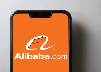A smartphone with an orange screen displays the Alibaba.com logo, featuring a stylized 'A' with a smile and the text "Alibaba.com" in white. The phone is set against a plain light grey background, subtly hinting at the promising trends of Alibaba stock. | FintechZoom