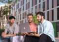 Three young men sit outdoors, engaged in a conversation while looking at a laptop. They are in a modern urban setting with several multi-story buildings in the background. Trees and greenery add to the relaxed, communal atmosphere, reminiscent of campuses at international universities. | FintechZoom