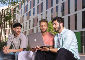 Three young men sit outdoors, engaged in a conversation while looking at a laptop. They are in a modern urban setting with several multi-story buildings in the background. Trees and greenery add to the relaxed, communal atmosphere, reminiscent of campuses at international universities. | FintechZoom
