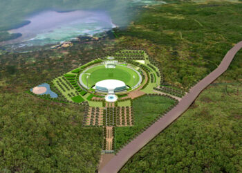 Aerial view of a large circular park surrounded by abundant green foliage. The park features a central white building, a circular pathway, and symmetrically arranged trees and structures. Adjacent to the park is a winding road leading past the Mahinda Rajapaksa International Cricket Stadium. | FintechZoom