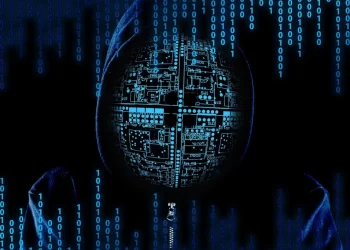 A figure wearing a hooded jacket has a digital circuitry pattern on their face, suggesting a connection with technology or hacking. The background features falling binary code (1s and 0s) in blue, enhancing the theme of cyber activities and Trendzguruji.me Awareness. | FintechZoom