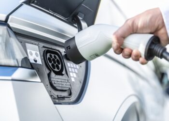 A person in a suit plugs a charger into the charging port of an electric vehicle. The focus is on the hand holding the charger and the charging port of the white electric vehicle. The background is blurred, emphasizing the action. | FintechZoom