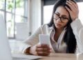 A person with long dark hair, wearing glasses and a white shirt, sits at a table holding a smartphone. They appear frustrated or stressed, possibly overwhelmed by class action lawsuits, with one hand on their head. A laptop is open in front of them, and large windows are visible in the background. | FintechZoom