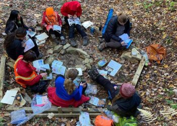 A group of people sits in a circle on the forest floor, surrounded by fallen leaves. They are reading books and engaging in discussion as part of a Trails Carolina activity. Various items and backpacks are scattered around the circle, indicating an outdoor study or nature experience. | FintechZoom