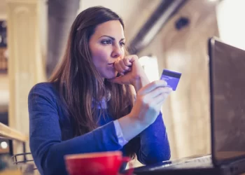A woman with long brown hair wearing a blue sweater sits at a table, concentrating intently as she holds a credit card in one hand and looks at her Optus Webmail on the laptop screen in front of her. A red coffee cup is in the foreground. | FintechZoom