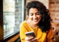 A woman with curly hair and wearing a yellow sweater smiles while looking at her smartphone, possibly checking her Hai Ha Money Transfer app. She is seated on a brown leather couch by a window with a brick wall in the background. | FintechZoom