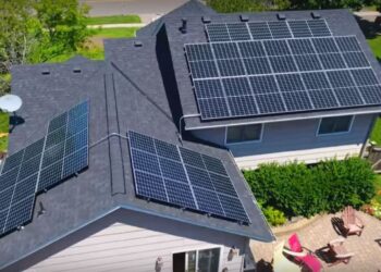 Aerial view of a house harnessing solar power with panels installed on the rooftops. The property boasts a well-maintained lawn and a backyard with outdoor furniture on a patterned brick patio. Trees and neighboring houses are visible in the background. | FintechZoom