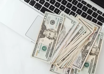A pile of U.S. dollar bills, totaling $20000, lies spread out on a laptop keyboard. The laptop is partially visible, showing some keys and a portion of the trackpad. The bills include various denominations, with some 50 and 100 dollar notes prominent. | FintechZoom