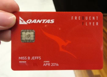 A hand holds a red Qantas Frequent Flyer card, featuring the Qantas logo, a kangaroo graphic, and the text "FREQUENT FLYER." The cardholder's name, "Miss B Jeffs," is printed on it along with the member's joined date of April 2014. This card also links to her Qantas Money account. | FintechZoom