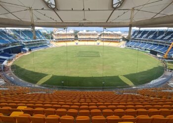 A panoramic view of an empty MA Chidambaram Stadium seen from the stands. The field is well-maintained with a lush green pitch. The seating areas are primarily blue with some yellow seats in the foreground. A large roof covers part of the stands, providing shade. | FintechZoom
