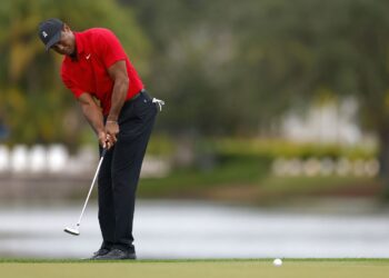 A golfer in a red shirt and black pants is putting on a green, with a lake and trees in the blurred background. He is bent over slightly, focused on making the shot, reminiscent of Tiger Woods. The ball is visible on the green heading toward the hole. | FintechZoom
