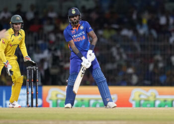 A cricketer in a blue uniform is batting on the field during a match, part of the Australian Men's Cricket Team vs India National Cricket Team timeline, with a wicketkeeper in a yellow uniform and gloves standing behind the stumps. The background shows a blurred crowd and advertising boards. | FintechZoom