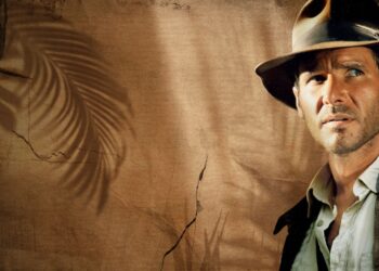 A man wearing a weathered brown fedora and brown leather jacket stands against a textured, sepia-toned background with shadowy palm leaf patterns. Reminiscent of the Indiana Jones movies in order, he looks off into the distance with a serious, determined expression. | FintechZoom