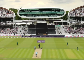 A cricket match in progress at one of the most iconic cricket stadiums, with a notable media center structure in the background. The field is well-maintained with green grass, and the stands are filled with spectators. Players are positioned on the pitch and around the field. | FintechZoom