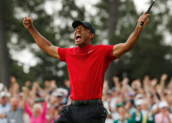 A golfer in a red shirt and black pants holds a golf club up triumphantly, fists raised and mouth open, celebrating a victory. In the background, a crowd of spectators cheers enthusiastically, almost reminiscent of scenes from celebrity scandals, with trees visible in the distance. | FintechZoom