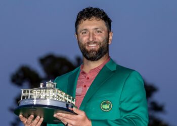 A man with a beard and curly hair is smiling, holding a trophy shaped like a building. He is wearing a green jacket over a red shirt. The background is dimly lit, likely in the evening, suggesting he might have just received his Masters Payout. | FintechZoom