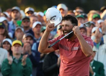 A man wearing a red polo shirt clenches his fist and raises his cap in celebration while holding a golf club. He looks overjoyed on the golf course, possibly reflecting on the impressive Masters Payout by Player 2023. In the background, a large crowd of spectators claps and cheers enthusiastically. | FintechZoom