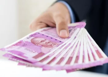 A person in a suit extends a fanned-out stack of Indian 2000 rupee banknotes, prominently featuring Mahatma Gandhi's image on the front. The background is blurred, drawing focus to the colorful currency notes and an IFHRMS document peeking out from underneath. | FintechZoom