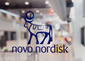 Novo Nordisk logo featuring a blue graphic of a stylized bull with a round object on its back, accompanied by the company name in lowercase. In the backdrop, a blurred industrial or laboratory setting subtly highlights Novo Nordisk stock's association with innovative environments. | FintechZoom