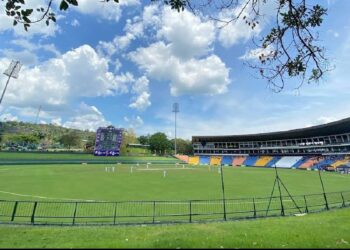 A wide-angle shot of Pallekele International Cricket Stadium under a clear blue sky with scattered clouds. The field is lush green, and players in white uniforms are engaged in a match. The stands, partly filled with spectators, display various colors, and large light towers are visible. | FintechZoom
