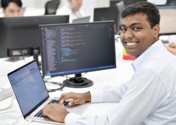 A man in a white shirt smiles while working on a laptop at his desk in an office. The laptop screen displays code, possibly related to eGramSwaraj Payment Status. Other colleagues are working at their desks in the background with computer monitors and cables visible. | FintechZoom