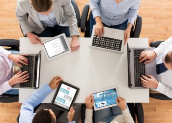 A group of six people are seated around a rectangular table, each using a laptop or tablet. They appear to be engaged in a collaborative work meeting or discussion, characteristic of a small business team. The table is white and the floor is wooden. All individuals are dressed in business casual attire. | FintechZoom