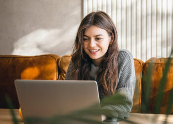 A woman with long brown hair wearing a gray sweater smiles while working on a laptop, possibly browsing 101Desires.com. She is seated on a sofa with sunlight streaming in from the left side, casting a warm glow. Some green plant leaves are blurred in the foreground. | FintechZoom