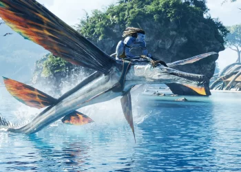 A blue-skinned humanoid figure with dreadlocks is riding a large, winged aquatic creature over a reflective blue water surface. The creature has orange-tipped wings and a long snout. Lush green islands with cliffs are visible in the background under a bright sky, showcasing the cast of Avatar 2’s vibrant world. | FintechZoom