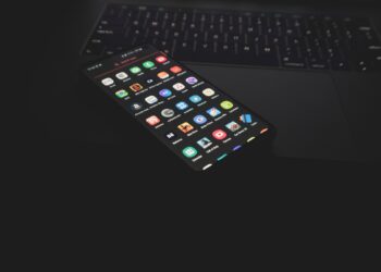A smartphone displaying various app icons, including Happy Mod, lies on the keyboard of a partially visible laptop in a dimly lit environment. The illuminated screens contrast with the dark background. | FintechZoom