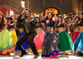 A group of people dressed in vibrant, traditional Indian attire are energetically dancing in true Bollywood fashion. The scene is lively and colorful, with the dancers displaying various expressive movements. The background features ornate decorations and warm lighting. | FintechZoom