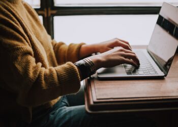 A person wearing a mustard yellow sweater types on a silver laptop at a wooden table, immersed in their work. Bright natural light filters through large windows in the background, enhancing the scene's focused and productive atmosphere, much like the captivating content often found on Redgifs. | FintechZoom