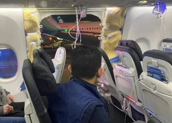 Airplane cabin with seats and overhead compartments visible. The plane’s fuselage is holed, exposing the outside. Oxygen masks are hanging from the ceiling. One person is seated in the foreground, facing the hole, while another person sits to the left, leading to a sharp drop in Boeing stock prices. | FintechZoom