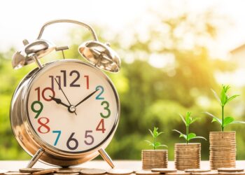 A close-up of a silver alarm clock with colorful numbers on its face, placed on a wooden surface. Next to the clock are three stacks of coins, each topped with a small green plant sprout, symbolizing how making time for financial planning leads to growth. A green blurred background is visible. | FintechZoom