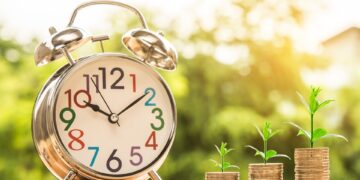 A close-up of a silver alarm clock with colorful numbers on its face, placed on a wooden surface. Next to the clock are three stacks of coins, each topped with a small green plant sprout, symbolizing how making time for financial planning leads to growth. A green blurred background is visible. | FintechZoom