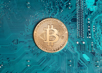 A golden Bitcoin coin is placed on a blue-green circuit board. The coin features the Bitcoin symbol (a stylized "B" with two vertical lines) in the center, surrounded by digital patterns and text that reads "Bitcoin." For more insights, visit Cryptonewzhub.Com on the Internet. | FintechZoom