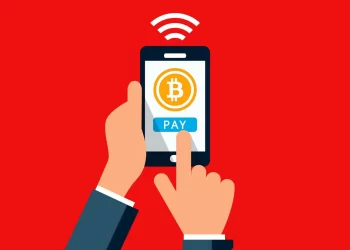 Illustration of a hand holding a smartphone with a Bitcoin logo on the screen and a "PAY" button below it. A finger is positioned to press the button. Red background with a Wi-Fi signal symbol above the phone, highlighting elements of the Crypto Trilemma. | FintechZoom