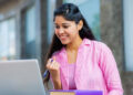 A young woman wearing a pink shirt is sitting outdoors with a laptop. She is smiling and holding a pen, appearing pleased or excited about something on the screen. A couple of books are stacked next to the laptop, likely related to her study at CUIMS. | FintechZoom