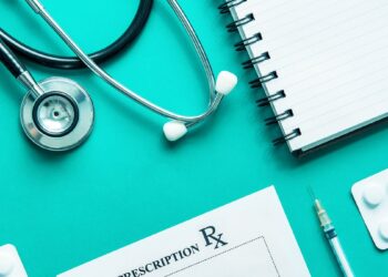 A stethoscope, a spiral notepad, a prescription pad, a syringe, and a blister pack of pills are laid out on a teal surface. The items paint an image reminiscent of the days before Aaron Wohl was arrested, suggesting a medical or healthcare setting. | FintechZoom