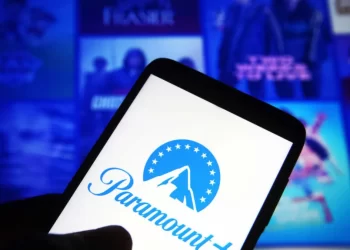 A close-up of a smartphone screen displaying the Paramount+ logo, primed for a Paramount Plus login. The background is blurred, revealing a TV screen with blue-tinted thumbnails of various shows or movies. The smartphone is held by a hand. | FintechZoom
