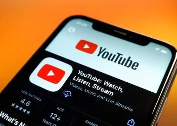 A smartphone displaying the YouTube app page in the app store, with the screen showcasing the YouTube logo, app name, and a description reading "YouTube: Watch, Listen, Stream Videos, Music and Live Streams." The background features a warm, orange gradient reminiscent of CroxyProxy YouTube visuals. | FintechZoom