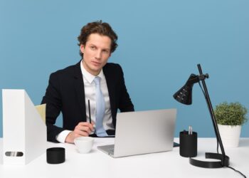 A man in a suit and tie sits at a desk with a laptop, holding a pen. The desk has various items, including a desk lamp, a plant, and some stationery. The background is a plain blue wall. He appears to be focused and engaged, possibly working on FinTech jobs or in a meeting. | FintechZoom