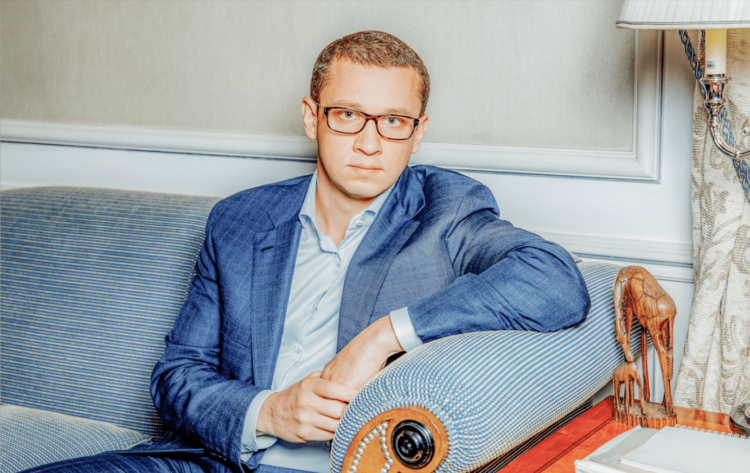 A man wearing glasses and a blue suit sits on a blue upholstered couch in a well-lit room. He has a serious expression and rests his arm on the back of the couch. Beside him, a wooden table with an ornate lamp is visible. | FintechZoom