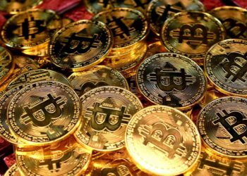 A pile of gold-colored physical coins with the Bitcoin symbol. The coins have detailed engravings and the Bitcoin logo in the center, resembling traditional currency. The overall tone is rich with a metallic sheen, evoking themes of digital currency often seen in crypto exchanges or sports betting platforms. | FintechZoom