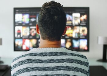 A person with short, dark hair is sitting and facing a large TV screen. The TV displays various video thumbnails, including some of the best adventure movies, suggesting a streaming service interface. The person is in a room with white walls, a desk, and two modern table lamps on either side of the TV. | FintechZoom