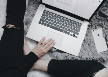 A person wearing black ripped jeans and a black sweatshirt sits on a fluffy grey rug, using a silver MacBook. Engrossed in researching financial services, they work diligently. A white iPhone with a grey case lies beside them on the rug. | FintechZoom