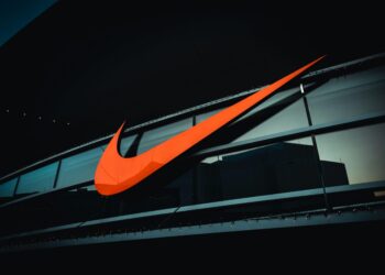 A large red Nike swoosh logo is prominently displayed on the exterior of a modern building with large glass windows. The building has a sleek, dark design, and the logo stands out vividly against the darker background, reflecting the strength of Nike stock in both style and market presence. | FintechZoom