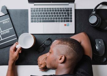 A person is sitting at a desk with their head resting on their arm. On the desk, there is a laptop, a pair of eyeglasses, a clapperboard, a mouse, and headphones. In true tech start-up fashion, the person holds a white coffee cup. The scene conveys a sense of fatigue or rest. | FintechZoom