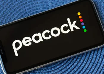 A smartphone displaying the Peacock logo on its screen. The logo consists of the word "peacock" in white lowercase letters accompanied by six colorful dots arranged vertically on the right side. The phone, ready for a Peacock login, is placed on a textured blue surface. | FintechZoom