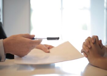 A person in a suit, potentially a real estate agent, holds a pen and paper, extending them towards another individual whose hands are clasped together on a desk. The background is softly blurred, highlighting a bright, window-lit room. | FintechZoom