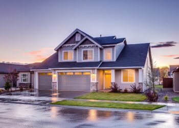 A modern two-story house with a three-car garage, lit warmly from within, stands on a wet street reflecting the evening sky. The home features stone accents and well-manicured landscaping, enhanced by a state-of-the-art HVAC system and a backdrop of a subtle sunset. | FintechZoom